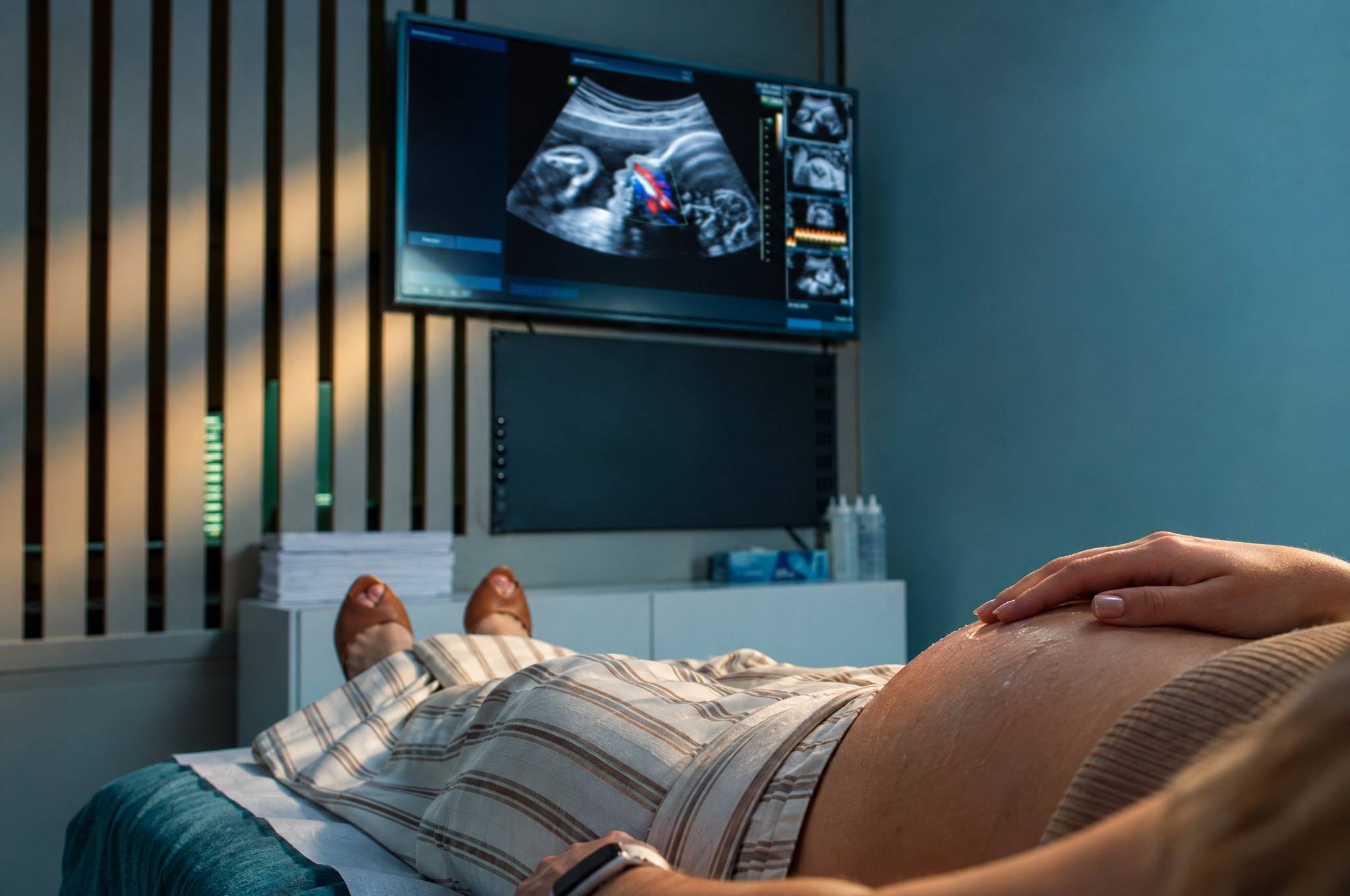 Gestante deitada para o exame, com a mão sobre a barriga, e o coração do bebê na tela ao fundo.