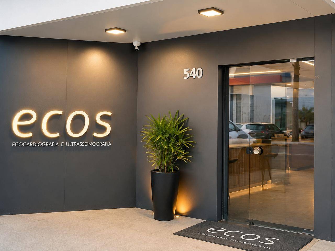 Fachada da Unidade I da ECOS: parede grafite com o letreiro "ecos" iluminado, número 540, planta e porta de vidro.